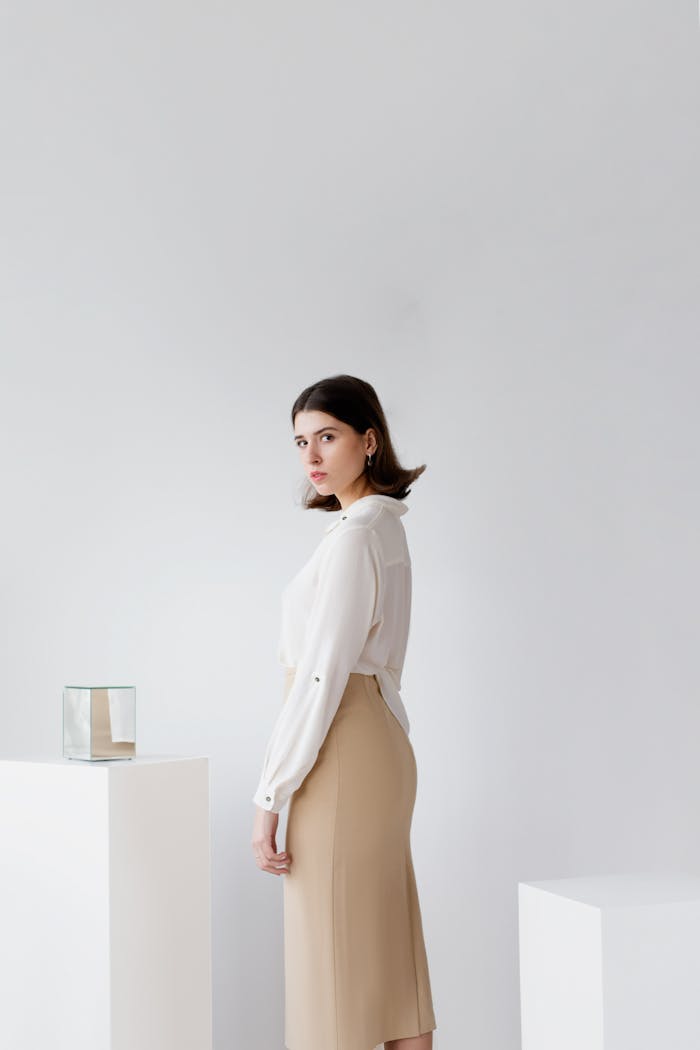 A fashionable woman poses indoors in a minimalist setting wearing a white blouse and beige skirt.