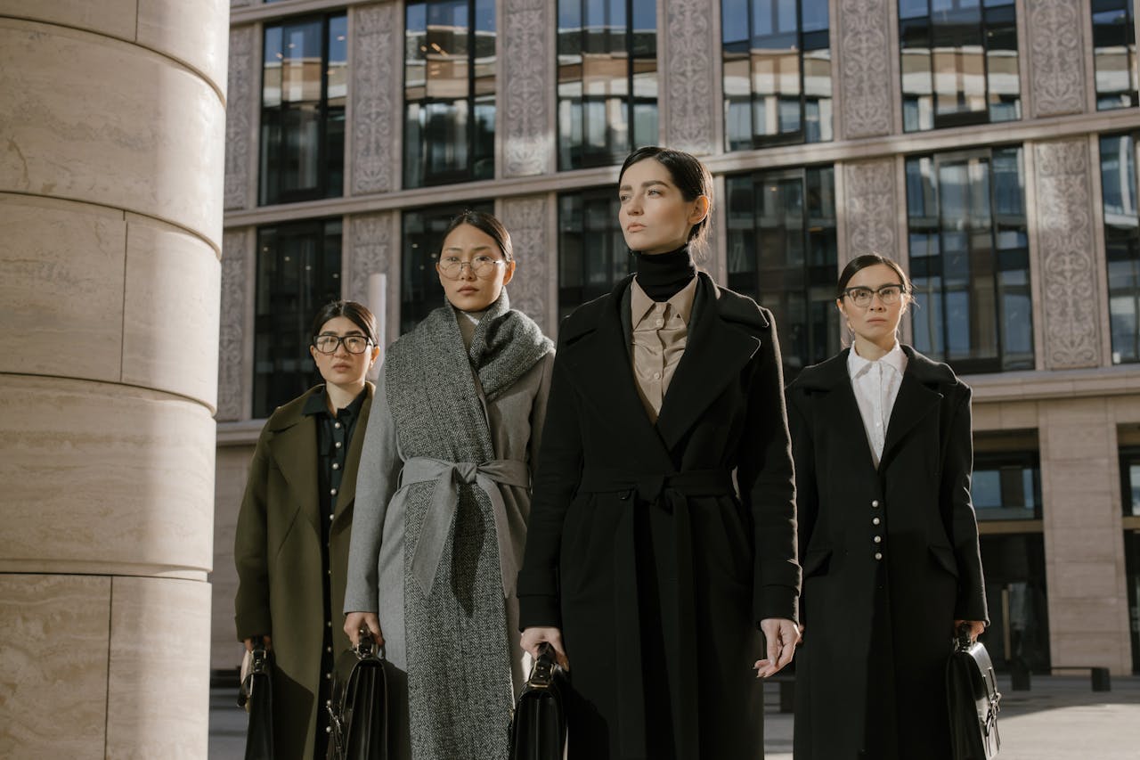 Four confident women in coats with briefcases outside an urban office building.